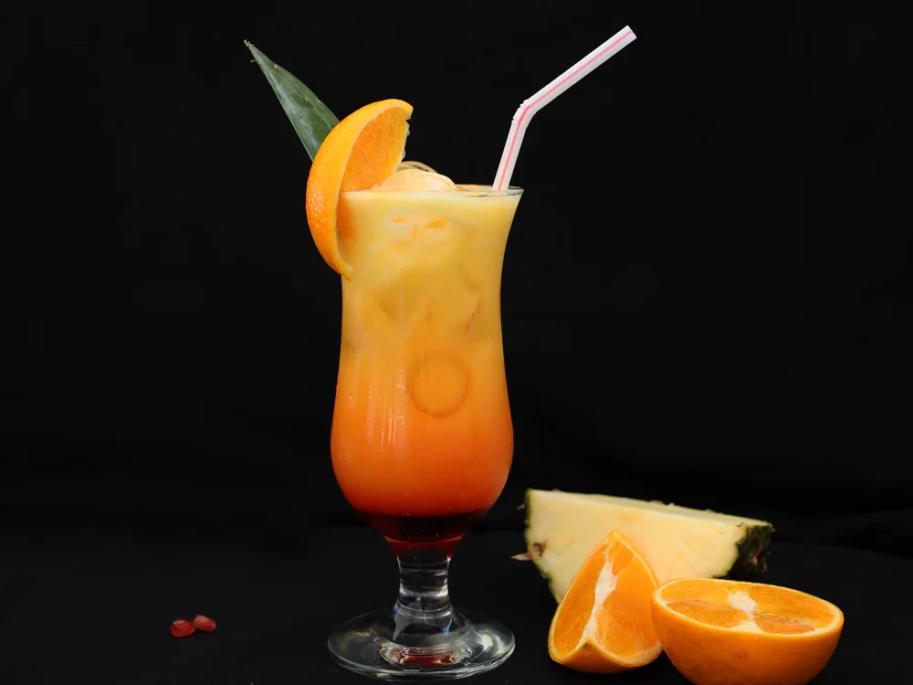 A tall tropical welcome drink garnished with orange and a pineapple leaf, with fresh pineapple and orange beside it
